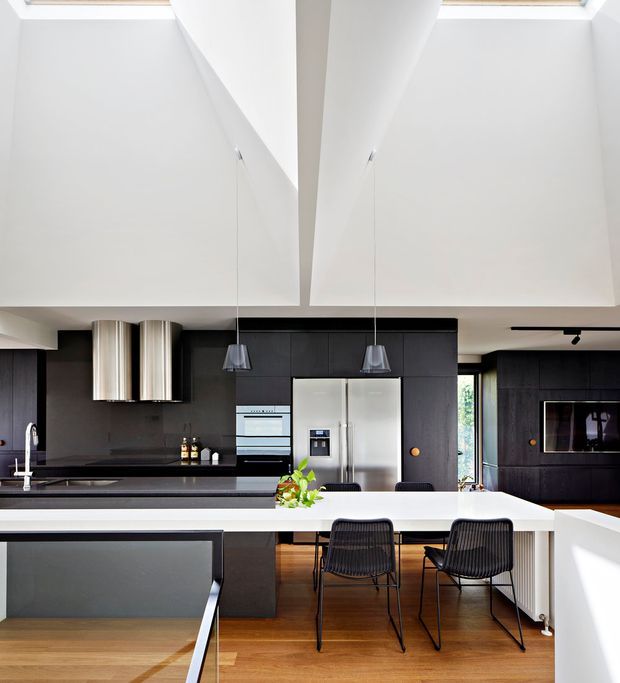 Bright contemporary kitchen with a large white island and integrated seating, dark charcoal cabinetry, stainless-steel refrigerator and appliances, black benchtops, pendant lights, and warm timber flooring. A dramatic sculptural white ceiling with high-level skylights creates an airy, double-height feel. The space includes a small dining area and built-in media cabinetry visible to the right.