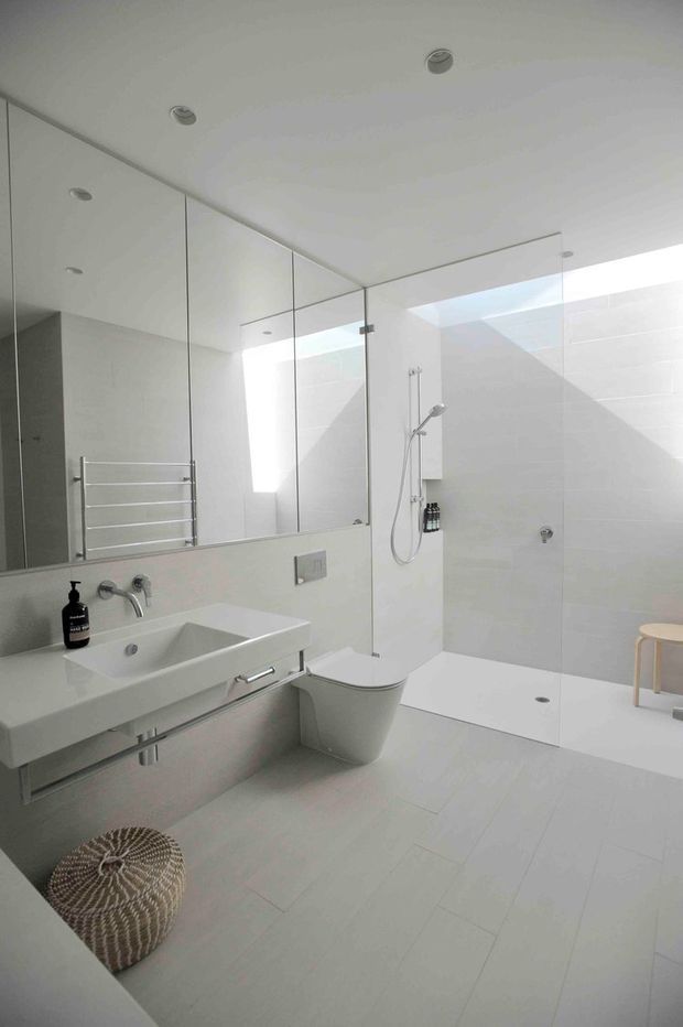 Bright, minimalist contemporary bathroom with a wall-mounted white basin and chrome tapware, floating vanity cabinetry, wall-hung toilet, large frameless glass walk-in shower, and a full-width mirrored wall. Pale large-format tiles and white surfaces create an airy, clean interior, with recessed ceiling lights and a narrow skylight or clerestory bringing in daylight.