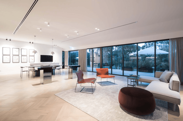 A bright, open-plan contemporary living and dining area with floor-to-ceiling glazing overlooking a landscaped outdoor space. The room features pale herringbone timber flooring, white walls and ceiling, a minimalist dining table with pendant lights, a wall-mounted television, sculptural lounge chairs, a large light sofa, and a dark rounded ottoman on a neutral rug.