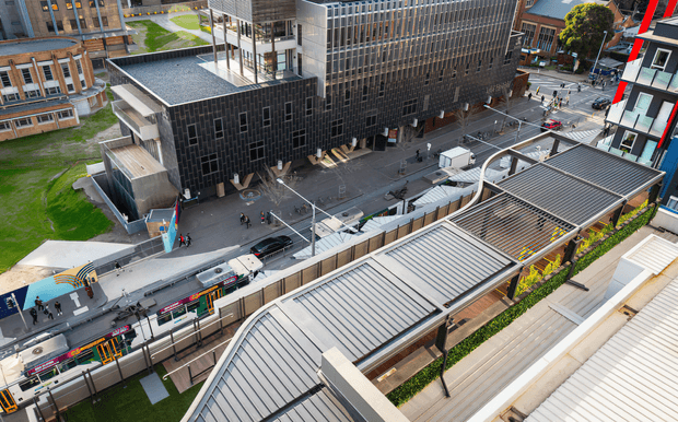 Elevated exterior view of a dense urban university or commercial precinct, showing contemporary multi-storey buildings with dark metal-clad façades, extensive glazing, covered pedestrian walkways, a street with trams, and layered ribbed metal roof structures in the foreground.