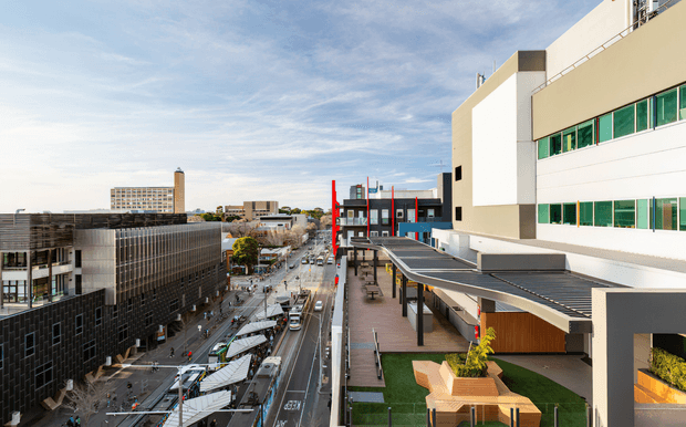 Elevated exterior view of a contemporary urban commercial or institutional precinct, looking along a city street with modern multi-storey buildings, covered pedestrian canopies, rooftop terraces, and landscaped outdoor seating. The main building on the right has a clean white and grey facade with long horizontal green-tinted window bands.