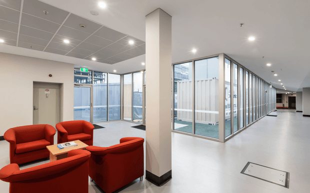 Bright contemporary commercial interior featuring a red upholstered seating area around a small table, a large square structural column, extensive internal glass partitioning, tiled suspended ceiling, recessed lighting, and a wide circulation corridor. An elevator and exit glazing are visible at the rear.