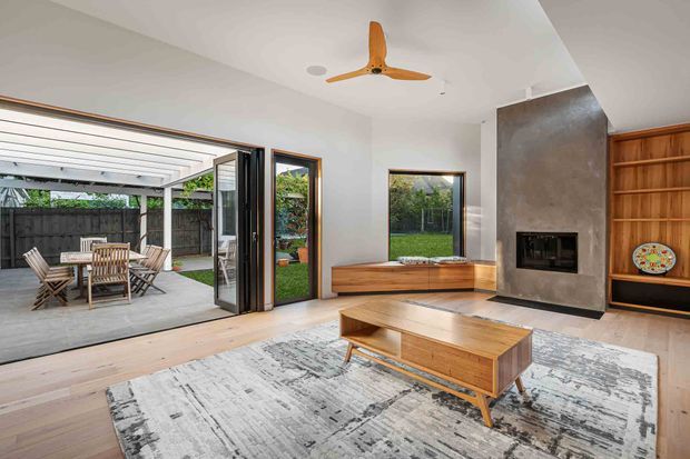 Contemporary open-plan living room with pale timber flooring, a large textured rug, and a low timber coffee table and built-in window seat. Full-height glazed doors open to a covered outdoor dining patio, while a dark stone or concrete fireplace surround and timber display shelving form a feature wall. A timber ceiling fan and abundant glazing complete the bright, indoor-outdoor space.