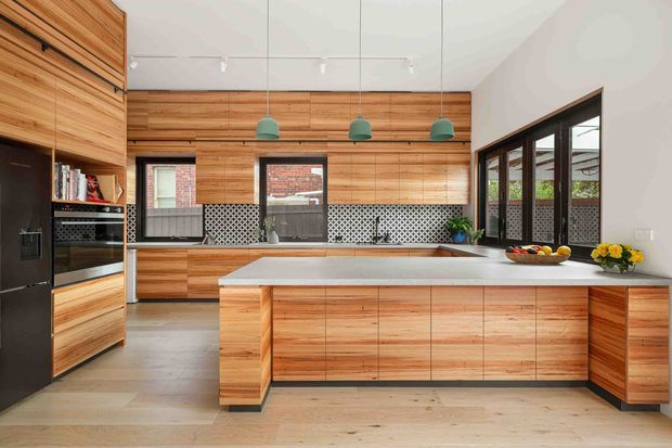 Bright contemporary kitchen featuring extensive warm timber cabinetry and wall panelling, a large pale stone island, dark appliances, patterned tile splashbacks, black-framed glazing, and three teal pendant lights. The kitchen opens toward an outdoor courtyard or garden visible through the windows.