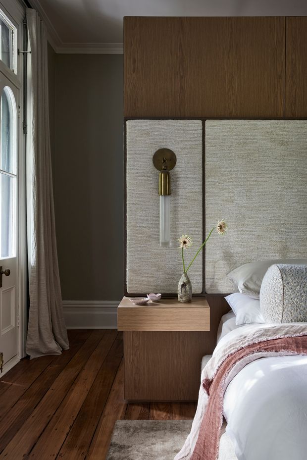 A refined bedroom interior featuring a bed beside a custom timber and upholstered feature wall. The built-in joinery has warm brown wood cabinetry above textured beige upholstered panels, with integrated brass-and-glass wall lighting and a floating timber bedside table. Dark timber floorboards, taupe walls, full-length curtains, layered white bedding, and a muted blush throw create a warm, sophisticated atmosphere.