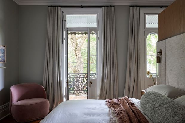 Elegant bedroom with tall French doors opening to a balcony, framed by full-height grey curtains. The room features a low upholstered bed with layered neutral bedding, a curved mauve armchair, dark grey walls, ornate balcony railings, and warm wood built-in joinery.
