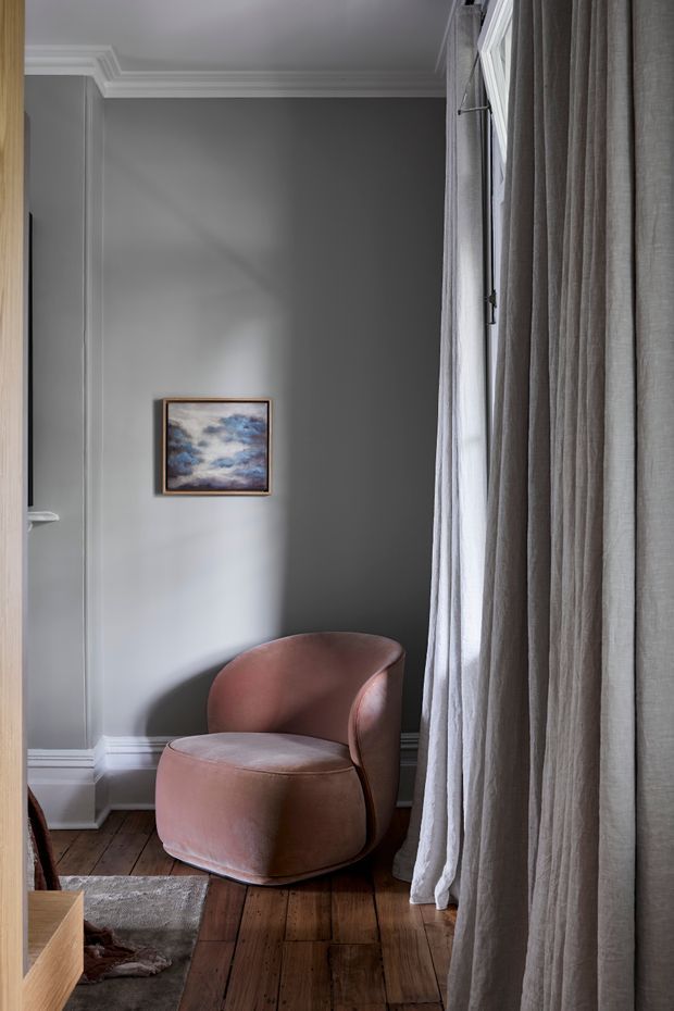 A refined interior corner featuring a rounded blush-pink upholstered lounge chair beside a tall window dressed with full-length textured grey curtains. The room has dark grey painted walls, white decorative crown moulding and skirting, aged timber floorboards, and a small framed artwork. Natural light creates a calm, softly layered atmosphere.
