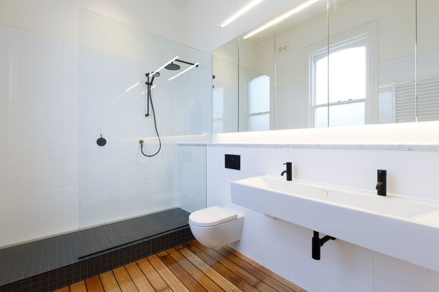 Bright contemporary bathroom with a walk-in shower enclosed by clear glass, a wall-hung toilet, and a long floating double vanity beneath a large mirrored wall. White tiled walls and sanitaryware are contrasted with black fixtures, while warm timber-look floorboards and dark shower floor tiles add texture.