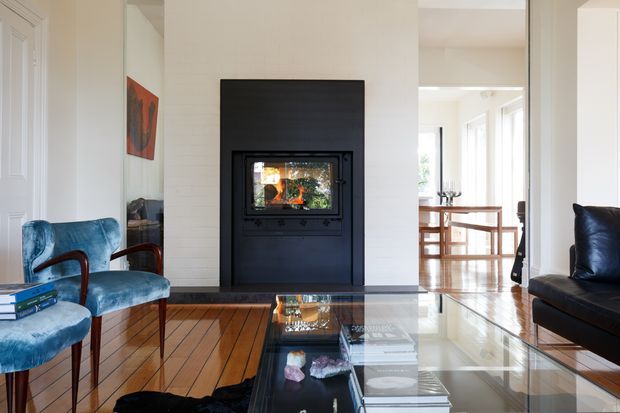 Bright, refined living room centred on a black built-in fireplace set into a pale painted masonry wall. The room has polished timber floorboards, a large glass coffee table, blue velvet occasional chairs, and a dark leather sofa. A glazed opening leads to a light-filled dining area with timber furniture and extensive windows.