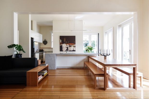 Bright open-plan interior combining a kitchen, dining area and living space. The room features polished timber flooring, a white kitchen with a large island, extensive glazing and doors, a black sofa, and a timber dining table with bench seating. A vertical panelled feature wall and artwork sit behind the island, with abundant natural light creating an airy contemporary atmosphere.