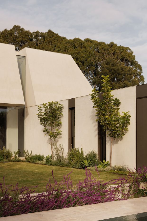 Exterior view of a contemporary minimalist residence with angular, sculptural pale rendered walls and narrow vertical glazing. The building is set within a landscaped garden featuring clipped lawn, ornamental trees, grasses, and purple flowering groundcover. A pale paved edge and a dark reflective strip appear in the foreground.