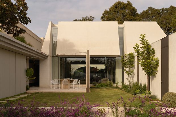 A refined contemporary residential courtyard facade with a minimalist cream-white geometric form, broad projecting upper volume, full-height glazed openings, and a sheltered outdoor dining terrace. The house is surrounded by manicured planting, lawn, and mature trees, with warm neutral tones and strong clean-lined composition.