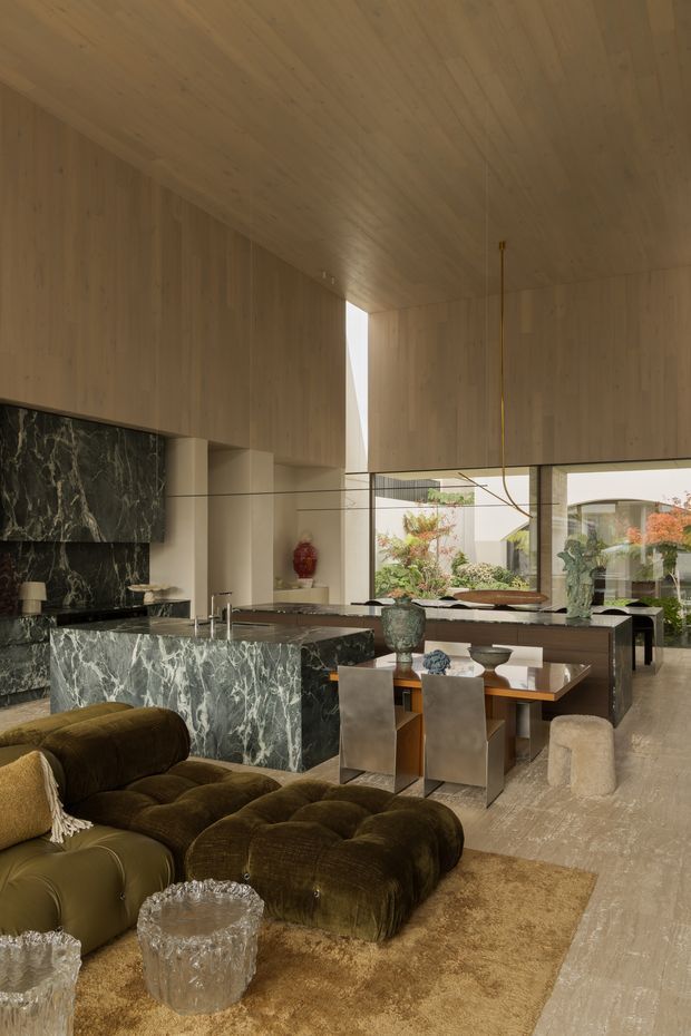 A dramatic open-plan interior combining living, dining and kitchen areas. Warm timber panels line the walls and ceiling, contrasting with a large dark veined stone kitchen island and fireplace surround. The room features an olive-brown tufted sectional sofa, a timber dining table with sculptural metal chairs, oversized glazed openings, sculptural decor and a muted neutral palette. The high ceiling and extensive glazing create a refined, luxurious atmosphere.