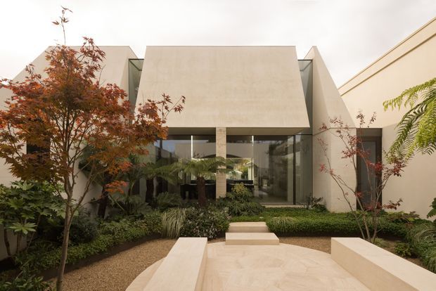 A contemporary single-storey residential facade viewed from a landscaped courtyard. The building has strong geometric forms, pale rendered walls, a projecting rectangular upper volume, extensive dark-framed glazing, and a sheltered entry approached by pale paved steps and a central walkway. Lush planting, ornamental trees, gravel beds, and integrated low seating walls frame the entrance.