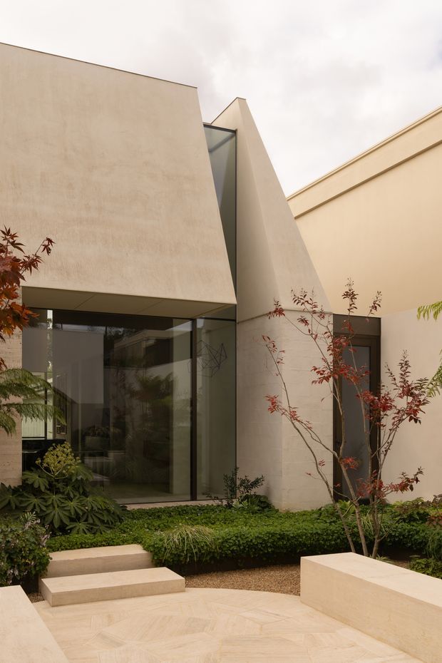 A minimalist contemporary courtyard garden viewed from outdoors, featuring sculptural cream-rendered geometric walls and angular roof forms, extensive dark-framed floor-to-ceiling glazing, pale stone paving and steps, raised cream masonry seating or planter walls, and layered greenery with small ornamental trees. The composition is calm and refined, with strong indoor-outdoor connection and subdued natural tones.