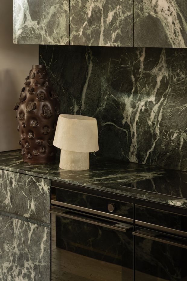 Close-up of a sophisticated dark green marble-clad built-in cabinet or sideboard, with dramatic pale veining across the benchtop, backsplash, cabinet fronts and upper panels. Black or very dark cabinetry includes integrated drawers and long metal pulls. A sculptural brown textured ceramic vase and small cream table lamp sit on the surface, creating a moody, luxurious interior vignette.