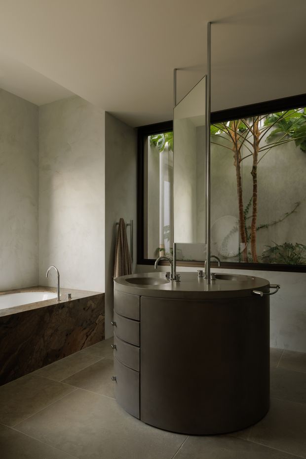 A moody, refined contemporary bathroom featuring a freestanding bathtub with a dark natural-stone or marble-clad surround, a large cylindrical double-sink vanity with integrated drawers, wall-mounted faucets, and a tall frameless mirror. Full-height black-framed glazing looks onto a planted courtyard, while muted plaster walls and large-format stone-look floor tiles create a calm, minimalist atmosphere.