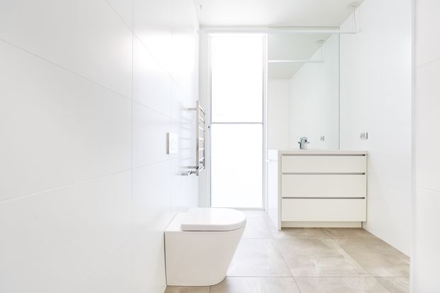 Bright minimalist bathroom with a wall-mounted vanity, integrated basin, large mirror, wall-hung toilet, heated towel rail, and pale large-format tiled surfaces. A tall frosted window provides soft natural light, creating a clean, airy feel.