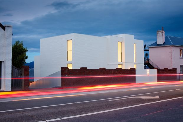 Twilight street-level exterior view of a crisp white contemporary multi-storey building with horizontal cladding, tall narrow illuminated windows, and a simple geometric form. A dark brick boundary wall separates the building from the roadway, where passing traffic creates vivid red and yellow light trails. Older neighbouring buildings and a dramatic blue sky frame the scene.