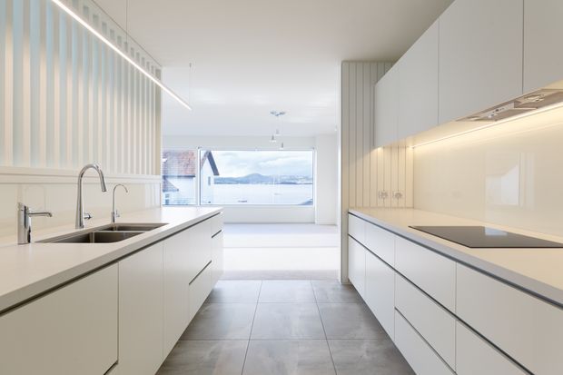 Bright, minimalist contemporary kitchen with a long central island, integrated sink and twin taps, extensive white flat-panel cabinetry, pale stone or engineered-stone benchtops, and a glossy light backsplash. Vertical fluted wall detailing and concealed linear lighting add texture. The kitchen opens toward a large glazed living or dining area with a distant water view.