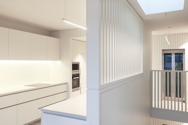 Bright minimalist open-plan interior showing a white kitchen with flat-panel cabinetry, integrated appliances and a pale benchtop, opening toward a hallway or landing. A vertical white slatted screen and matching balustrade define the adjacent stair void, with a large skylight and linear pendant lighting providing abundant natural light.