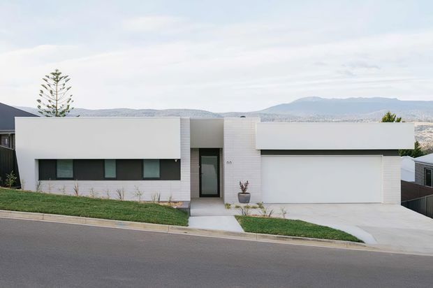 Street-facing view of a contemporary single-storey residence with a clean white rendered facade, long dark horizontal windows, a recessed entry door, and an integrated double garage. The house sits on a gently sloping suburban site with simple lawn strips and distant hills visible beyond.