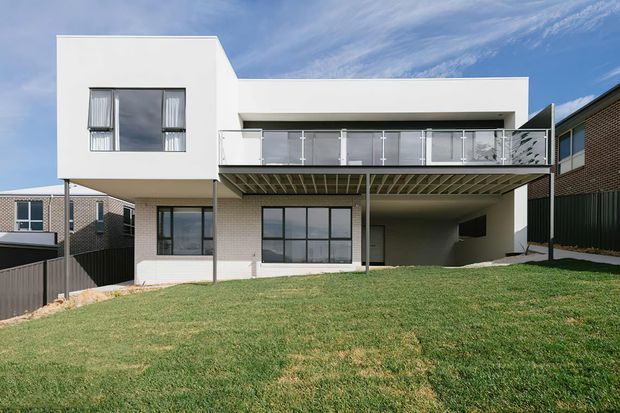 Rear exterior view of a contemporary two-storey residence with a white rendered cubic upper level, pale brick lower walls, dark-framed glazing, and a large elevated balcony with clear glass balustrades. The home opens onto a gently sloping lawn, with a covered ground-level area beneath the balcony and neighbouring suburban houses visible at the sides.