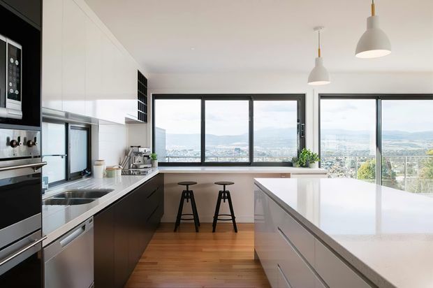 Bright contemporary galley kitchen with a long white stone island and integrated cabinetry, dark lower cabinets, timber flooring, stainless-steel appliances, and a breakfast bar positioned in front of expansive black-framed windows. The windows provide a panoramic view over a suburban landscape and distant hills. Two white pendant lights hang above the island.