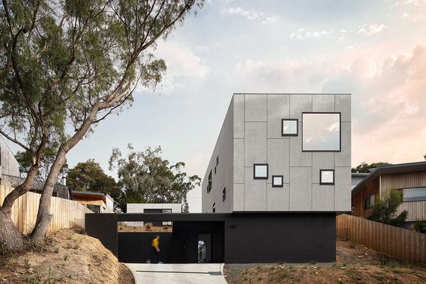 Exterior view of a contemporary multi-level residence on a sloped suburban site. The building features a rectilinear form, a dark ground-level base, a tall light-grey panelled upper volume with asymmetrically arranged square windows, and timber-clad neighbouring forms. Native trees and exposed earth frame the approach and reinforce the sloping setting.