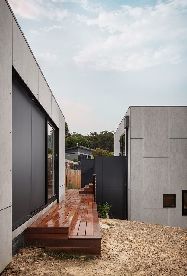 Exterior architectural view of a contemporary low-rise residence composed of two simple rectilinear volumes. Pale grey panel-clad walls, dark metal-framed glazing and doors, and a wet timber walkway and deck frame a narrow passage between the buildings. The scene includes a dark privacy gate, timber fencing, gravel landscaping, and a wooded suburban backdrop under a bright overcast sky.