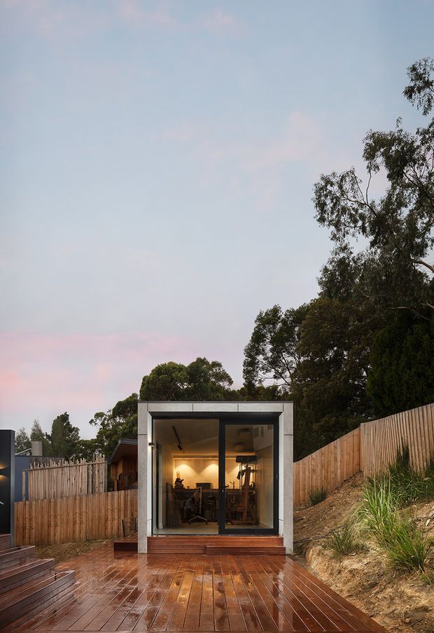 Exterior view of a compact modern garden studio or backyard workspace set into a sloping, wooded site. The structure has a simple box-like form with a flat roof, dark-framed floor-to-ceiling glazing, and light grey concrete or rendered framing. Warm interior lighting reveals a desk and workspace inside. A wet timber deck extends in front, bordered by timber fencing, planted banks, and mature trees under a soft evening sky.
