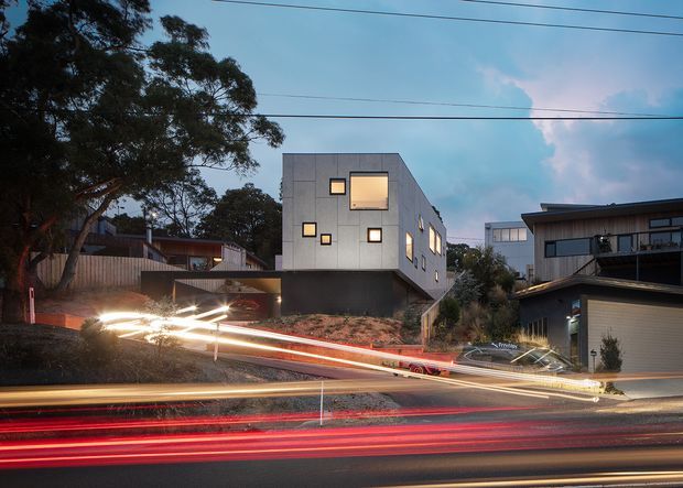 Dusk exterior architectural photograph of a contemporary multi-storey residential building on a sloping suburban site. The boxy grey-clad building has a dark lower level, asymmetrically arranged illuminated windows, and a flat roof. Adjacent modern homes, mature trees, overhead utility lines, and long-exposure vehicle light trails on the street are visible.