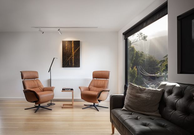 A bright, minimalist living room with two tan leather lounge chairs, a black tufted leather sofa, a slim floor lamp, and a small timber-and-metal side table. Light timber flooring and white walls create a clean backdrop, with a dark abstract artwork above a wall-mounted radiator. Large black-framed glazing opens the room visually to leafy surroundings, while a television is partially visible on the right wall.