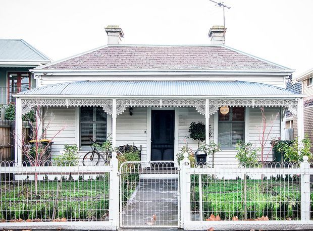 Front-on view of a compact heritage cottage with a symmetrical weatherboard facade, hipped roof, two chimneys, corrugated metal verandah roof, ornate lacework verandah detailing, timber-framed windows, and a small fenced garden with a central pedestrian gate. The building has a strongly historic Australian cottage character.