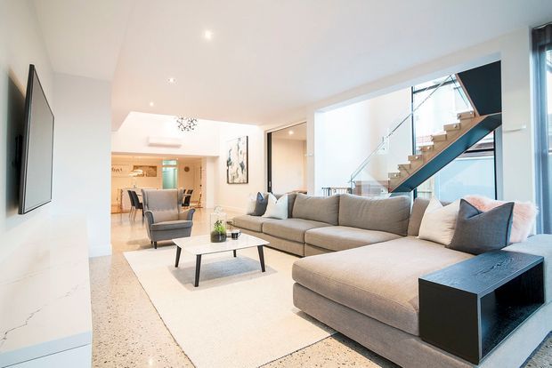 Bright contemporary open-plan living room with a large L-shaped grey sectional sofa, low black coffee table, pale area rug, wall-mounted television and minimalist decor. A dining area and kitchen are visible in the background, while a glass-balustraded staircase forms a prominent architectural feature along the right side. The space has white walls and ceiling, light terrazzo flooring, expansive glazing and a clean, refined finish.