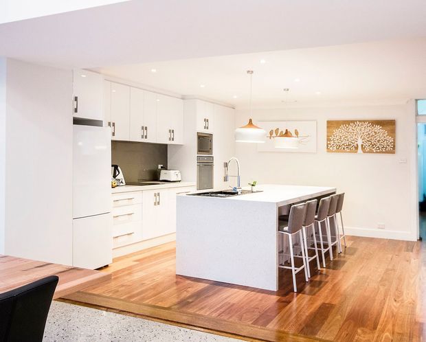 Bright, open-plan contemporary kitchen with white full-height cabinetry, integrated appliances, a large white island with sink and seating for four, timber flooring, and recessed ceiling lighting. A dining table is partially visible in the foreground, with simple framed artwork and a pendant light adding restrained decorative detail.