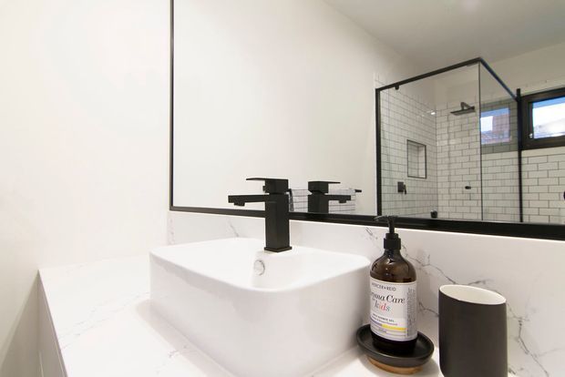A modern bathroom vanity with a white vessel basin on a white marble-look countertop, matte black faucet and fixtures, and a large black-framed mirror. The mirror reflects a walk-in shower enclosed by black-framed glass with white subway-tile walls and a small window. Minimal accessories sit beside the basin.