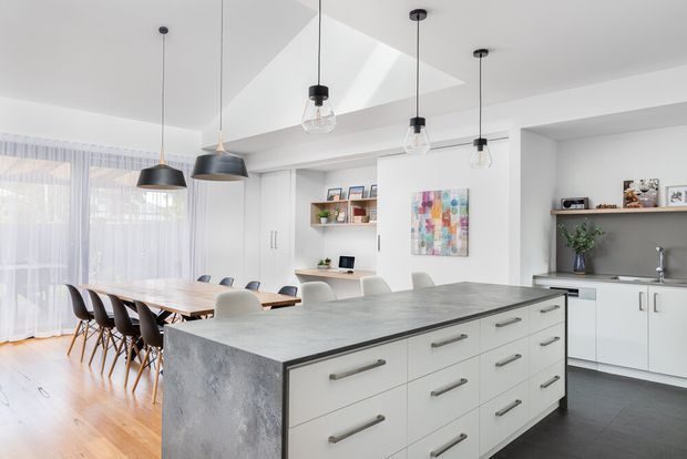 Bright open-plan kitchen and dining area with a large central island, white flat-panel cabinetry, grey stone-look benchtops, timber flooring, a timber dining table, and multiple pendant lights. Full-height sheer curtains line the dining area, while open shelving and a sink run are visible at the rear. A large skylight adds natural light.