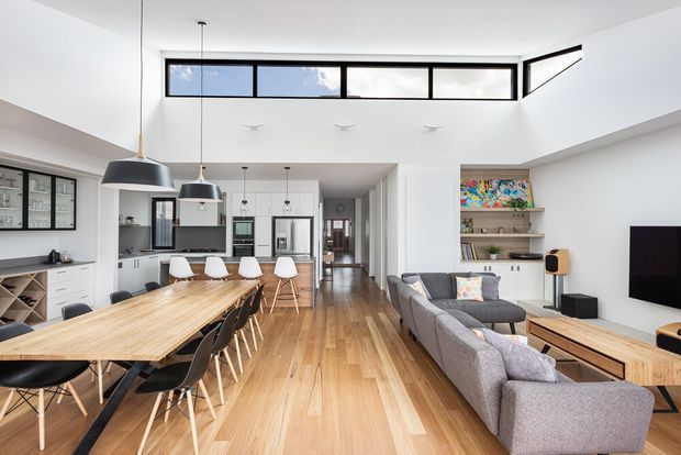 Bright open-plan interior combining a kitchen, dining area and living room beneath a high clerestory-lit ceiling. The space features warm timber flooring, a long timber dining table with dark dining chairs, a white kitchen with island seating, grey sectional sofa seating, pendant lights, built-in shelving and a wall-mounted television. Black-framed windows and doors provide strong graphic accents, while the upper glazing and white surfaces create an airy contemporary feel.