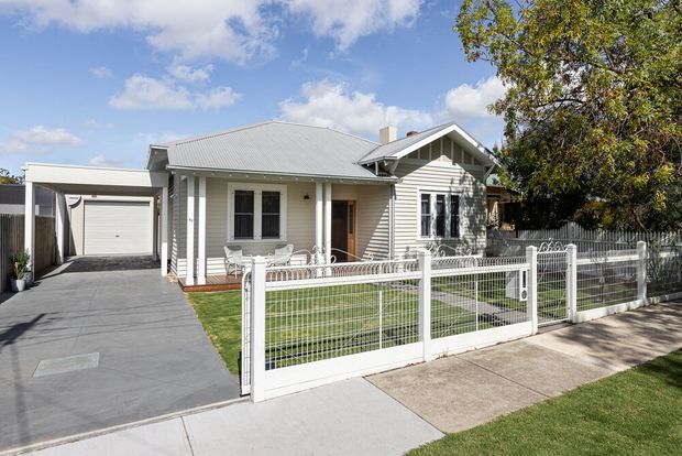 Street-facing exterior view of a single-storey suburban house with a light weatherboard facade, corrugated metal gabled roof, covered front porch, attached garage, paved driveway, and a white metal picket-style front fence enclosing a small lawn.