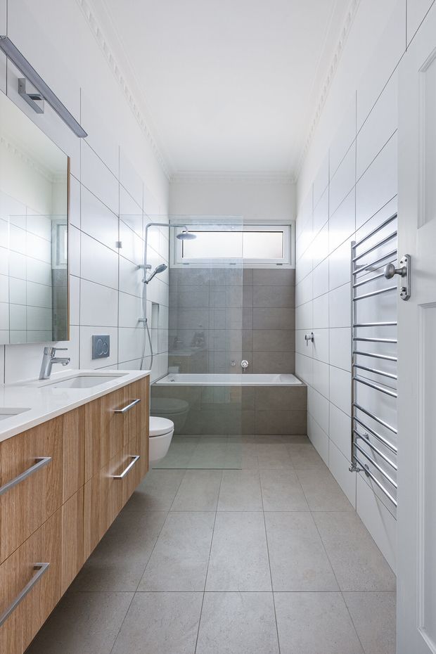 Bright, narrow contemporary bathroom viewed from the doorway. The room features large white wall tiles, light grey floor and bath-surround tiles, a timber-veneer floating vanity with white basin, mirrored cabinet, wall-hung toilet, glass shower screen, bathtub, chrome tapware, and a chrome heated towel rail. A high horizontal window provides natural light, while white decorative ceiling cornices add a subtle traditional detail.