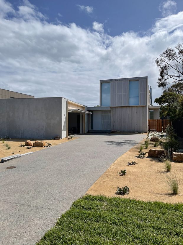 Exterior view of a contemporary two-storey residence with a flat-roofed, box-like form. The facade combines pale grey vertical cladding and corrugated metal screening with large blue-grey glazed openings. A broad paved driveway leads to an attached garage or carport, while the front landscape uses gravel, sandy planting beds, rocks and sparse drought-tolerant vegetation. Timber fencing and mature trees sit to the side under a bright, partly cloudy sky.