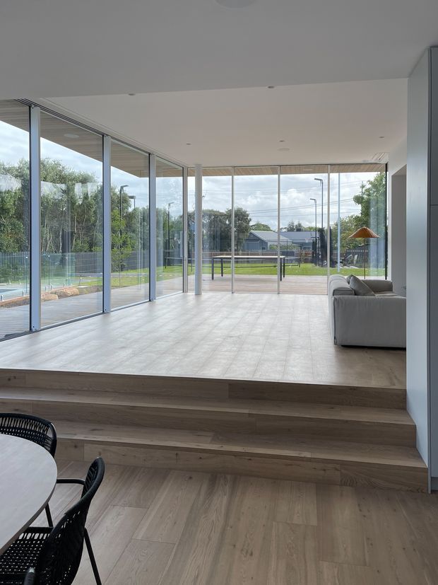 A bright, minimalist open-plan living and dining area with broad floor-to-ceiling glazing on multiple sides, opening onto a fenced landscaped outdoor area. The room has pale timber flooring, matching timber steps, a white ceiling with recessed downlights, slim dark window frames, and a light neutral sofa. A dining table and black woven chairs are partly visible in the foreground.