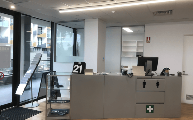 A modern commercial reception or service desk area with a large grey counter, computer workstation, queue number display, and drawers marked with male and female restroom symbols and a first-aid symbol. The space has full-height glazed frontage, white walls, a suspended grid ceiling with linear LED lighting, light timber-look flooring, and a small product display cabinet.