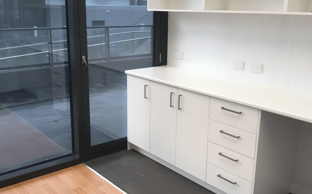 Compact modern kitchenette or utility joinery with white handle-front cabinetry, a pale solid benchtop, dark tiled flooring, and large black-framed glazed doors opening to an exterior balcony or walkway. The space is bright and highly functional, with clean contemporary finishes and minimal decoration.
