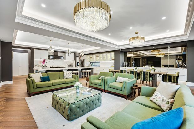 Open-plan luxury living area with several pale green upholstered sofas and armchairs arranged around a tufted ottoman coffee table on a light rug. The room opens to a white kitchen and bar area, with dark charcoal structural cabinetry or wall elements, warm timber flooring, layered coffered ceiling details, recessed lighting, and multiple crystal-and-gold chandeliers.