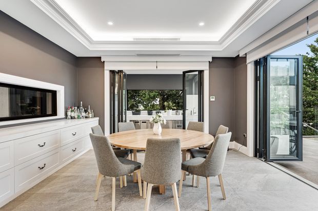 Refined dining room with a large round timber dining table and upholstered chairs, set within a bright contemporary interior. The room features dark taupe walls, white built-in cabinetry, elaborate white ceiling cornices and recessed lighting, large glazed doors opening to an outdoor terrace, and a view through to a kitchen or outdoor entertaining area.