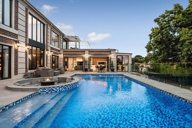 A large outdoor swimming pool dominates the foreground beside a multi-level brick-and-glass residence. The pool includes tiled steps, a raised circular spa with a small fire feature, and a glass safety fence. Wide paved terraces, outdoor seating, and a covered entertaining area connect the house to the pool, with mature trees beyond.