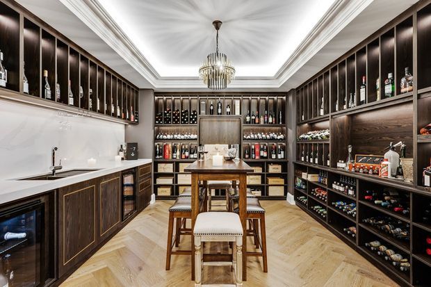 A luxurious, symmetrical wine room and entertaining space with dark timber built-in cabinetry lining three walls, illuminated bottle displays, a long central island with seating, a sink and under-counter wine refrigerator, and a coffered ceiling with a crystal chandelier. The herringbone timber floor and brass detailing create a refined, warm atmosphere.