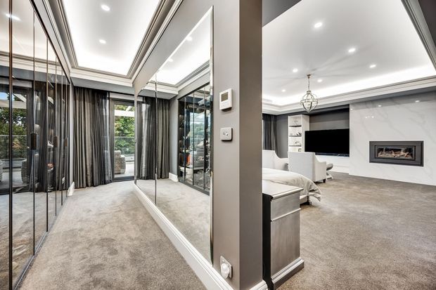 A refined contemporary interior showing a carpeted living room or media room opening to a mirrored hallway or dressing area. The space features extensive full-height mirrors, grey carpet, dark full-height curtains, recessed ceiling lighting, white built-in cabinetry, a wall-mounted television, and a contemporary recessed fireplace with a pale stone or plaster surround.